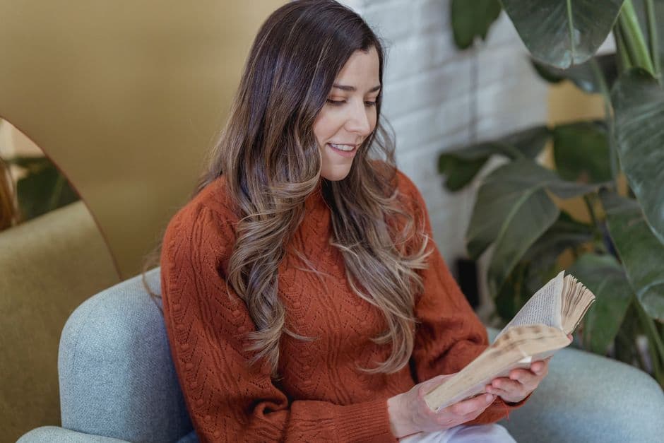 Cheerful female with long brown hair in casual clothes sitting on comfortable armchair and reading book in room in daytime