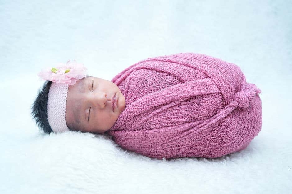 A peaceful newborn baby girl wrapped in a pink knitted blanket with a floral headband.