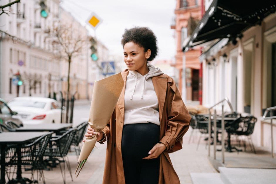 A stylish pregnant woman with afro hair holding flowers, walking down a charming city street.