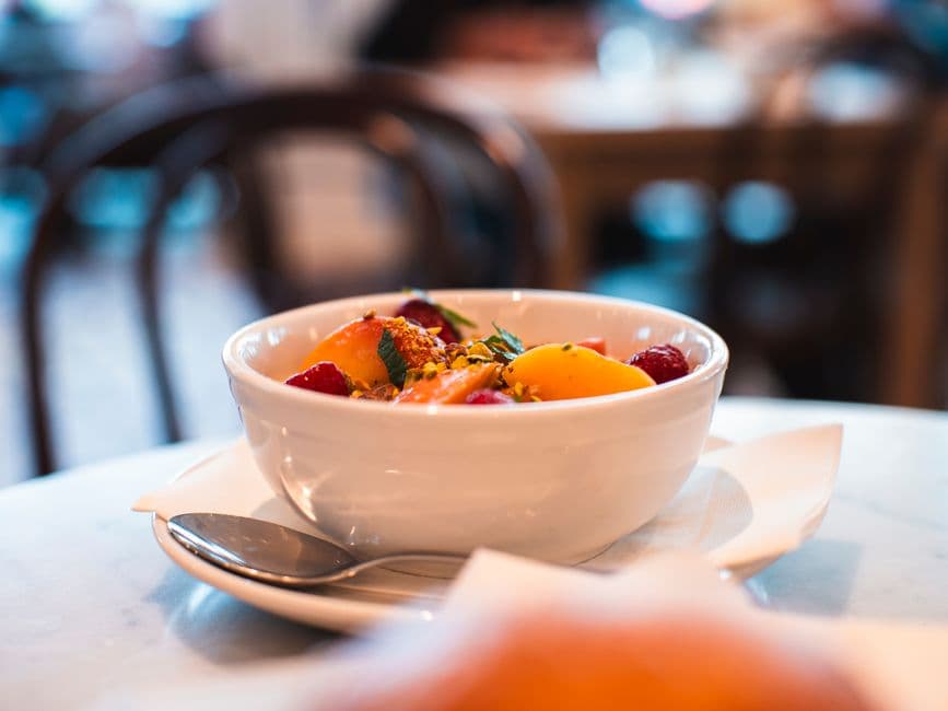 Close-up of a fresh fruit salad in a white bowl on a café table, brightly lit and inviting.