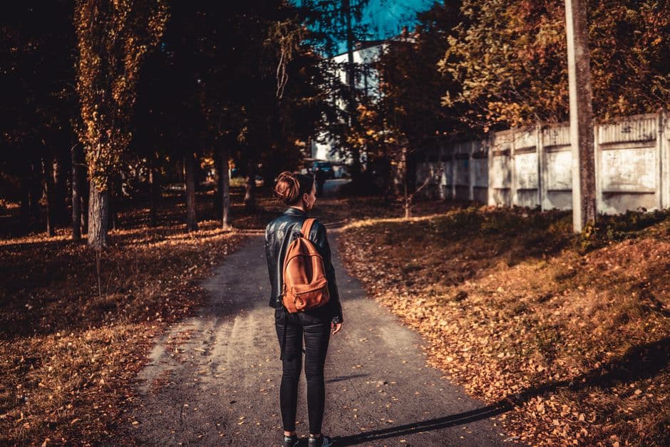 A woman with a brown backpack walks down a leafy path in the fall.
