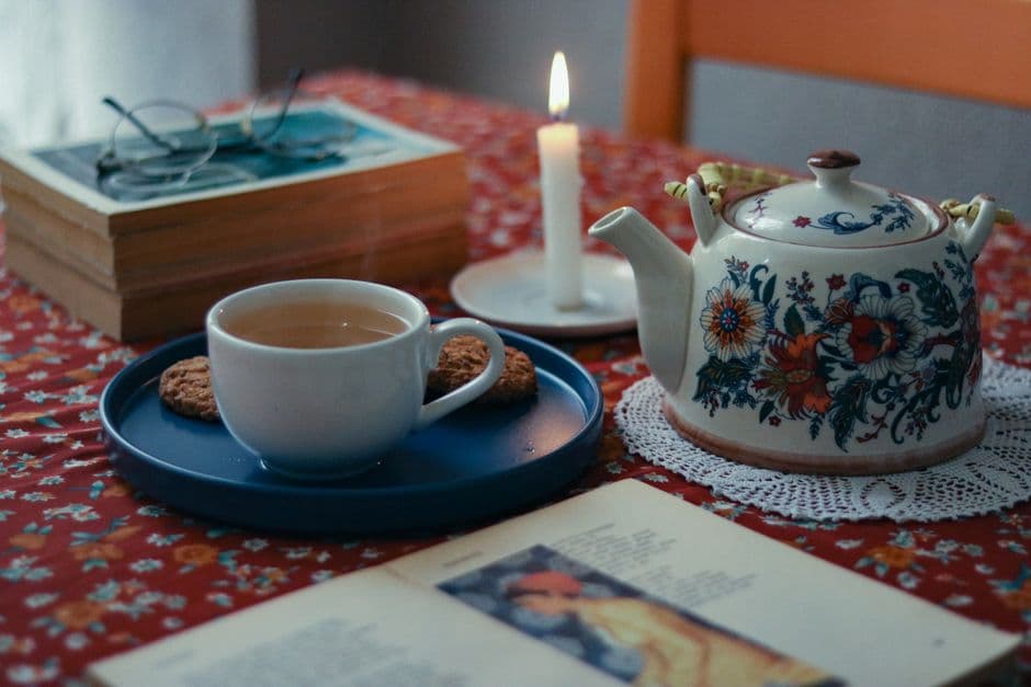 Warm indoor scene featuring a teapot, cup of tea, books, and a candle on a colorful tablecloth.