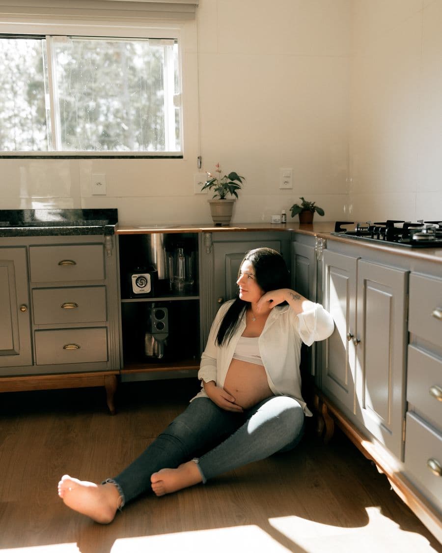 A pregnant woman sitting on a kitchen floor in afternoon sunlight, one hand resting on her bump