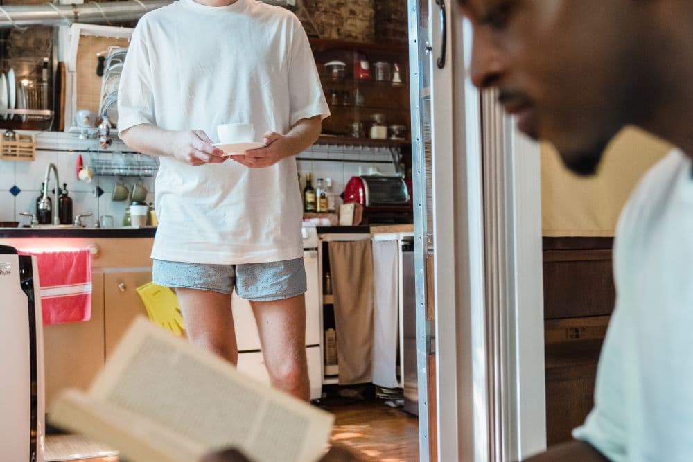 Two people sharing a quiet morning in a brick-walled kitchen, one reading and one carrying coffee