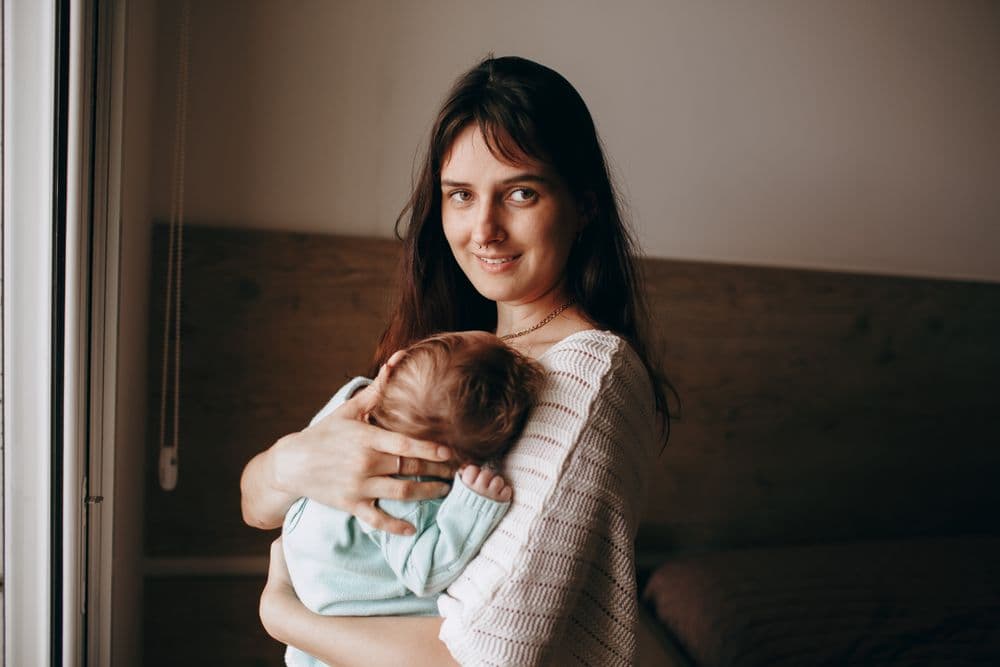 A parent holding a sleeping newborn beside a bedroom window