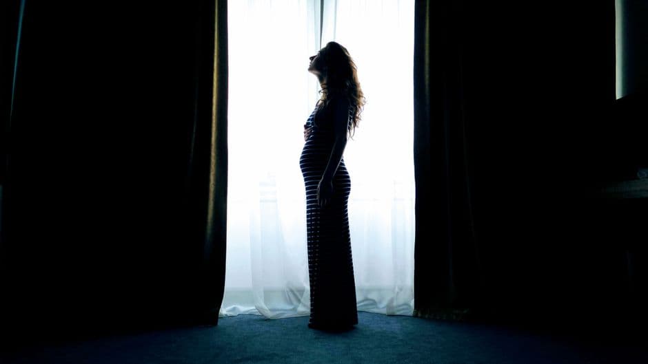 Silhouette of a pregnant woman standing by a bright window, captured in a serene indoor setting.