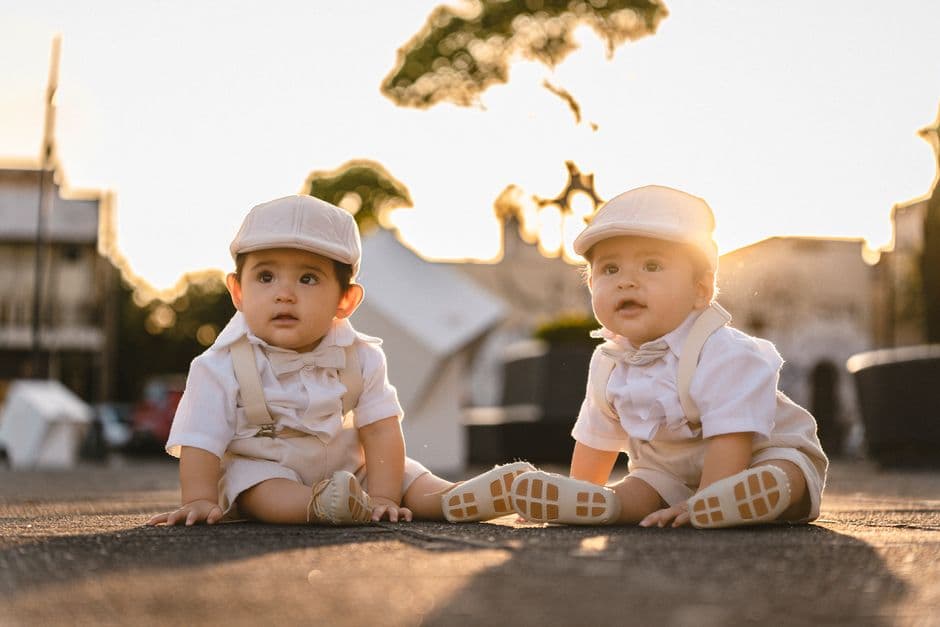 Adorable twin babies dressed in elegant attire sitting outdoors at sunset, creating a warm and cute scene.